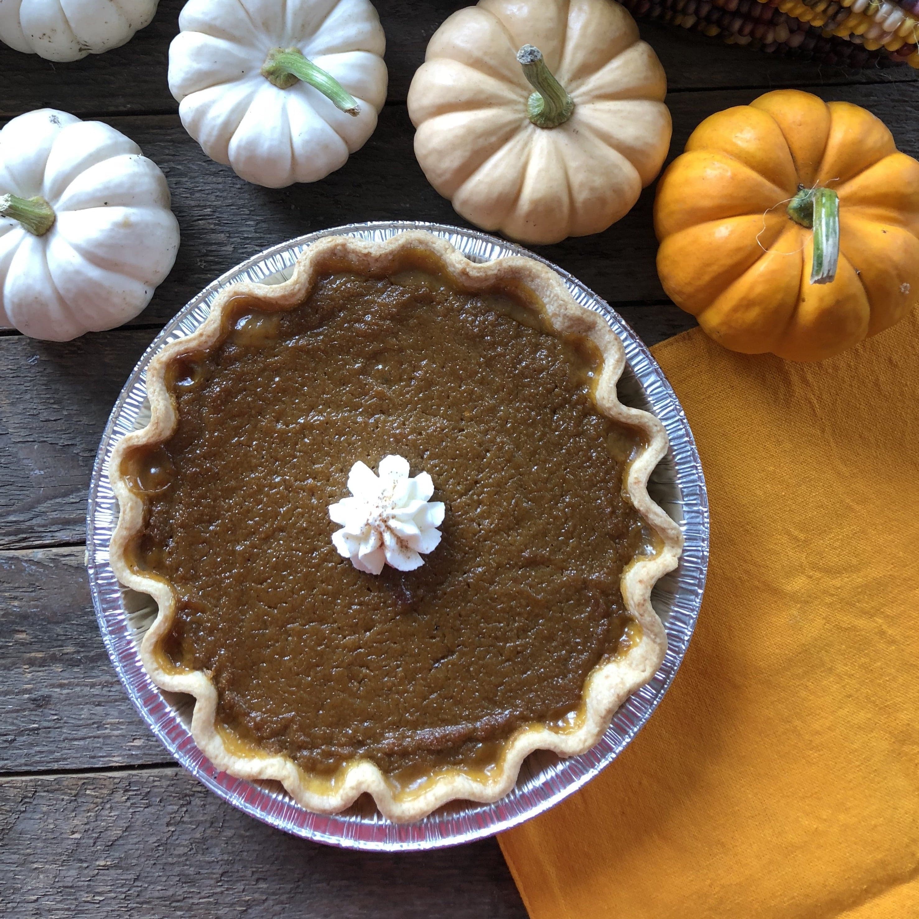 A pumpkin pie with a rosette of whipped cream on a weathered wooden table, ringed by small white and orange pumpkins and an orange napkin