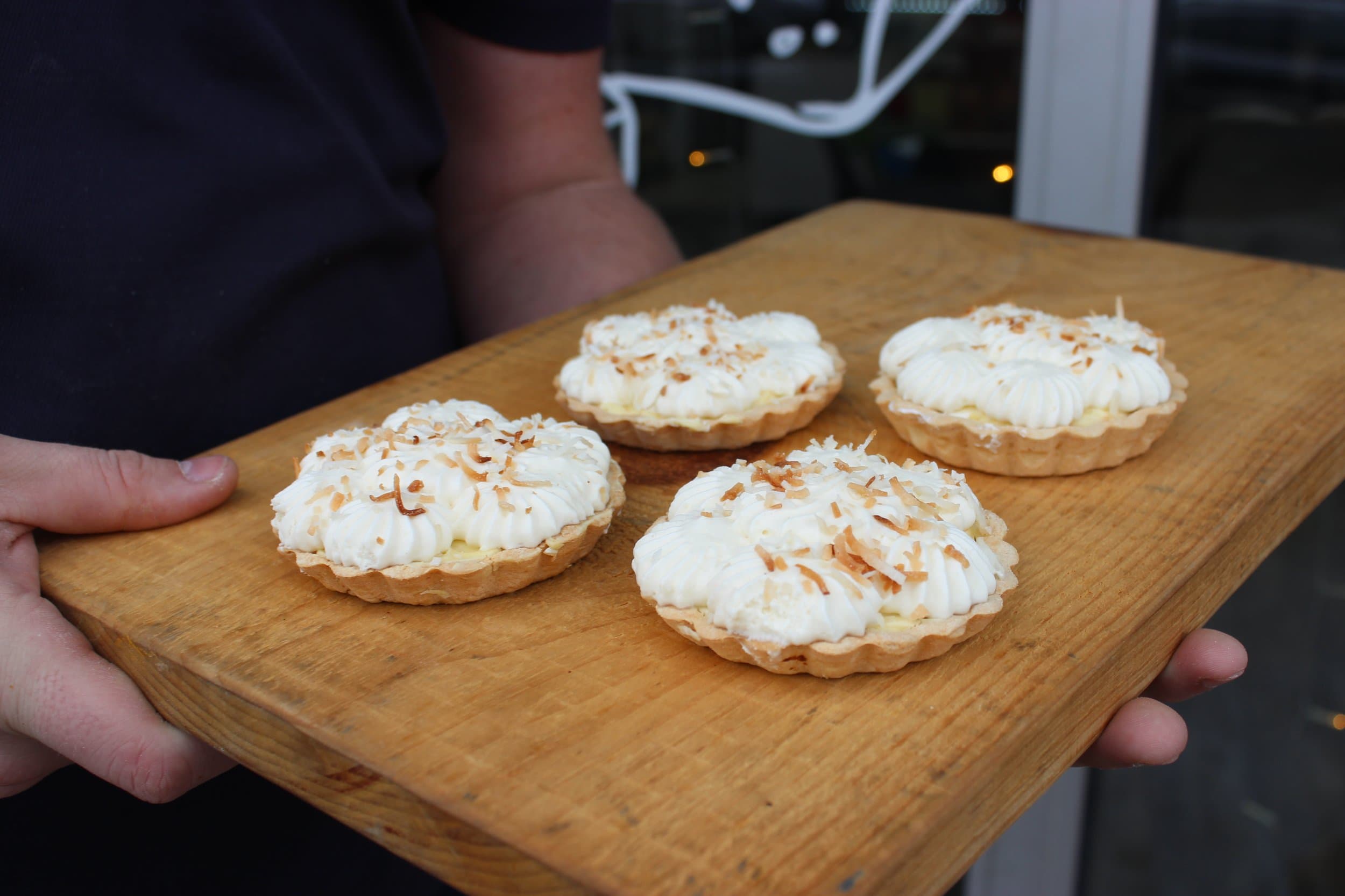 Four coconut cream tarts topped with piped cream and toasted coconut on a wooden board held out in two hands