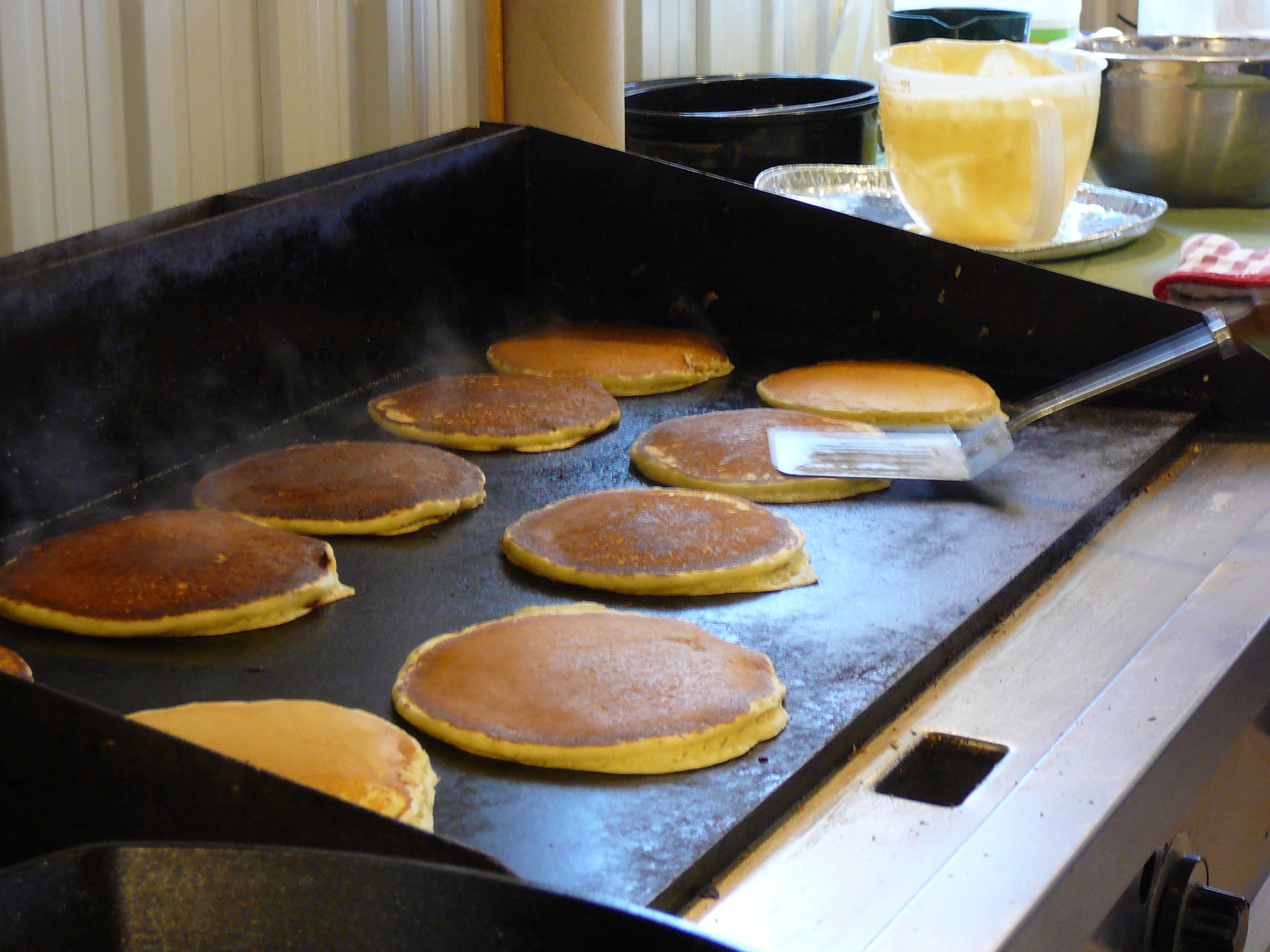 Pancakes cooking on a large black griddle, with a spatula lifting one and a jug of batter behind