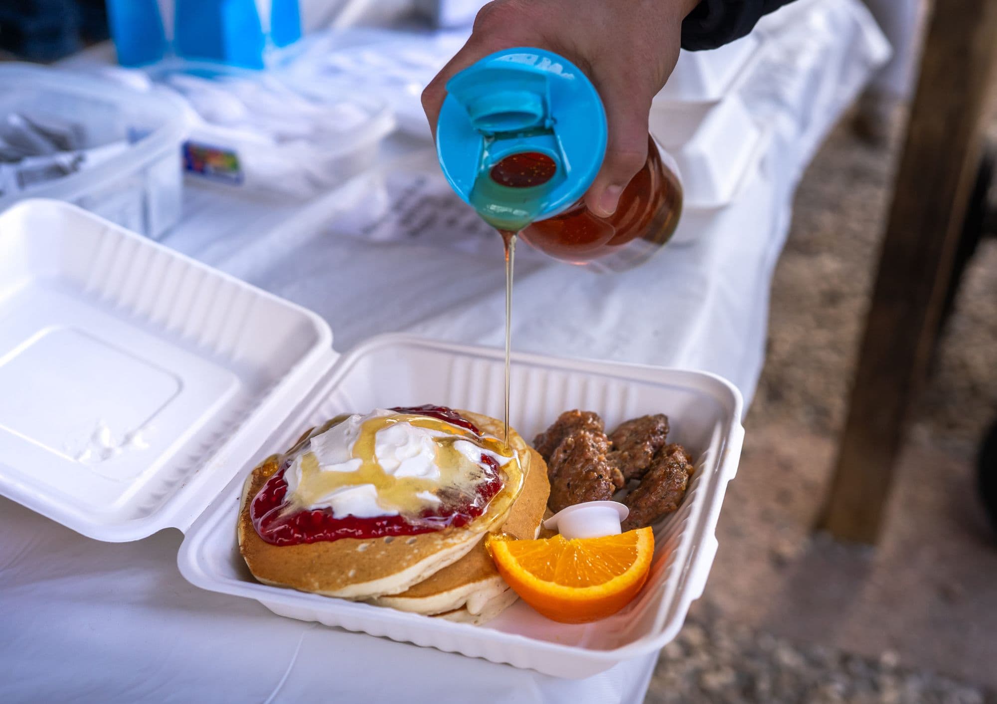 Maple syrup poured over pancakes topped with whipped cream and red berry sauce, with sausages and an orange slice in a takeout box