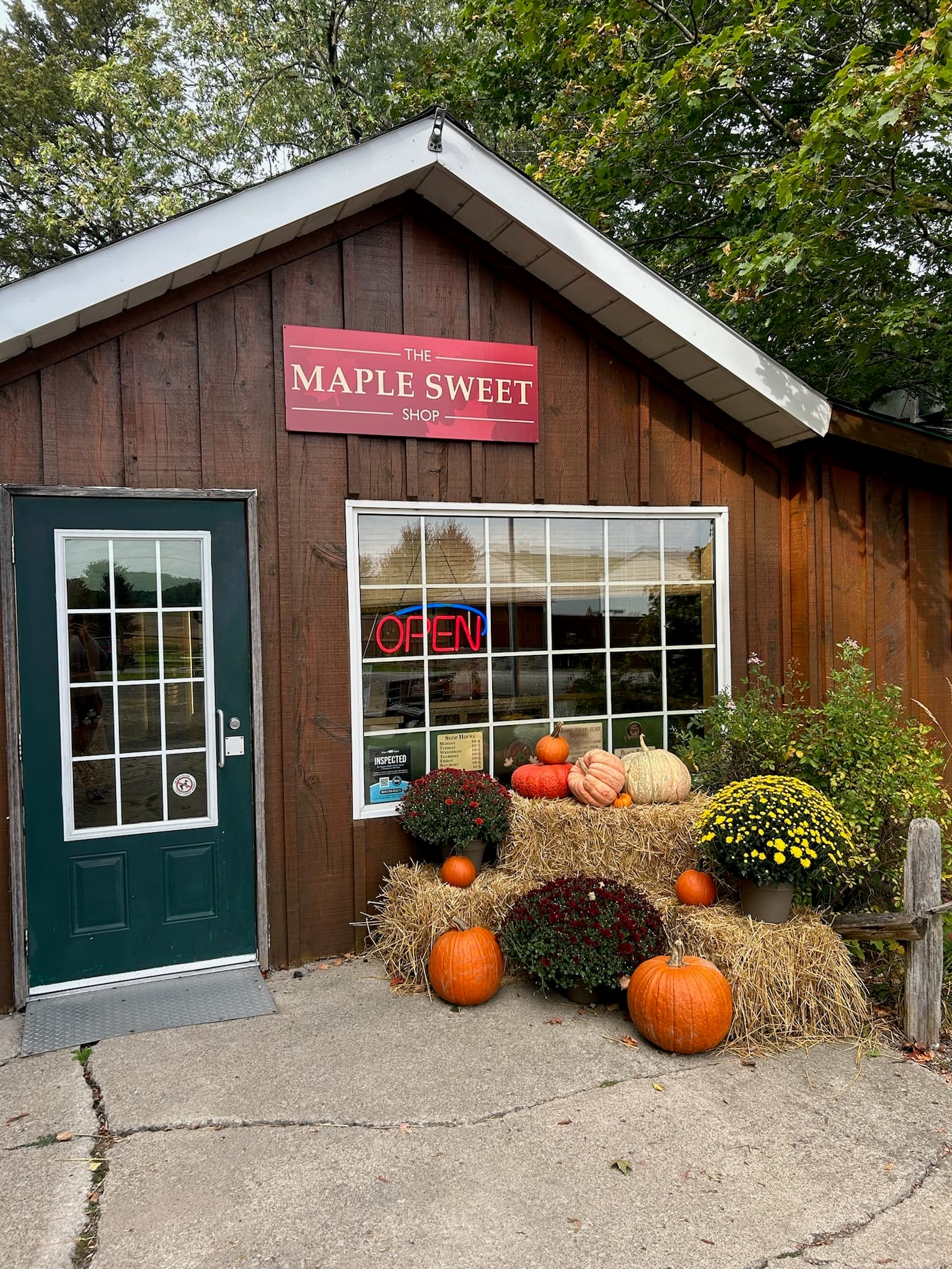 The Maple Sweet Shop's red sign over a brown board-and-batten shopfront, with pumpkins, potted mums and straw bales below the window