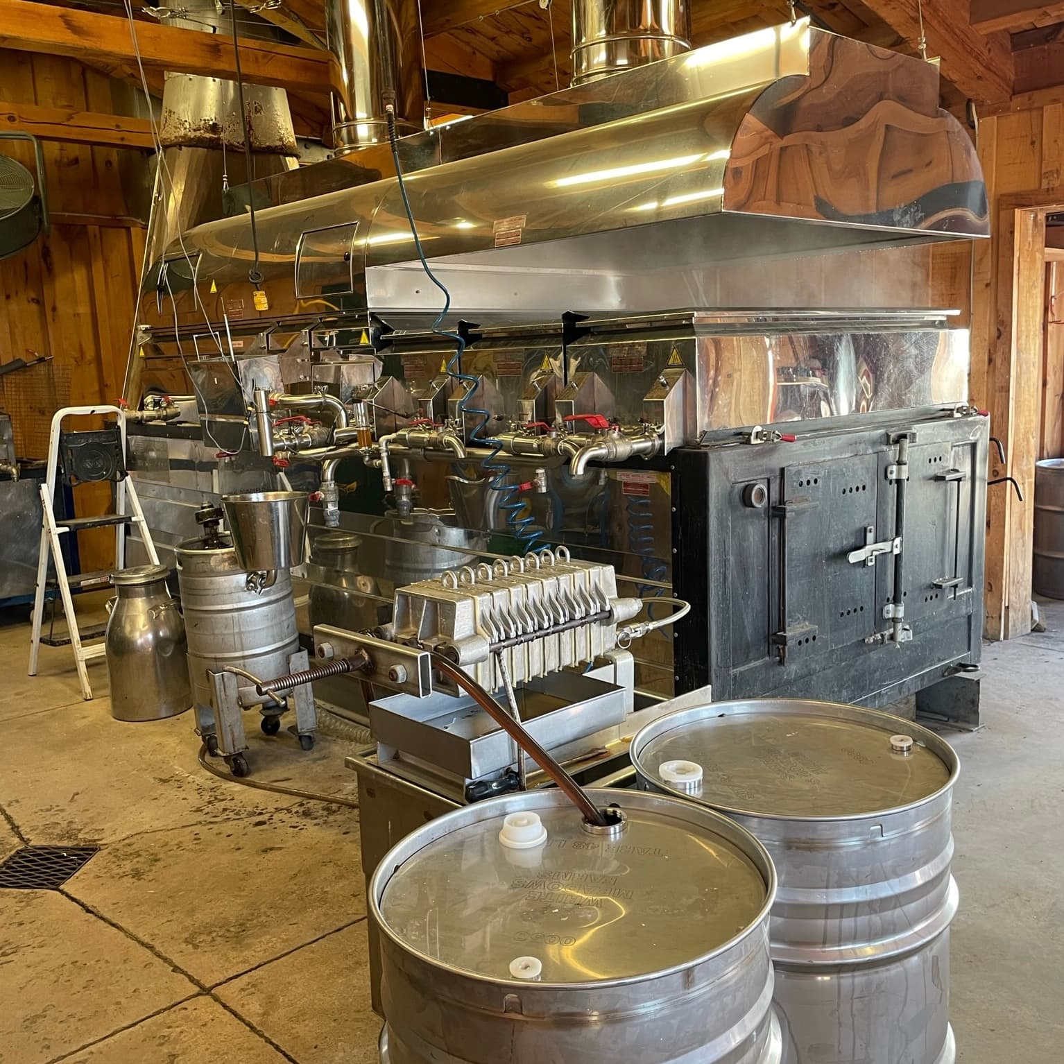 The evaporator in the Sugar Shack: a long stainless steel pan and hood over a black firebox, with a filter press and two steel syrup drums in front