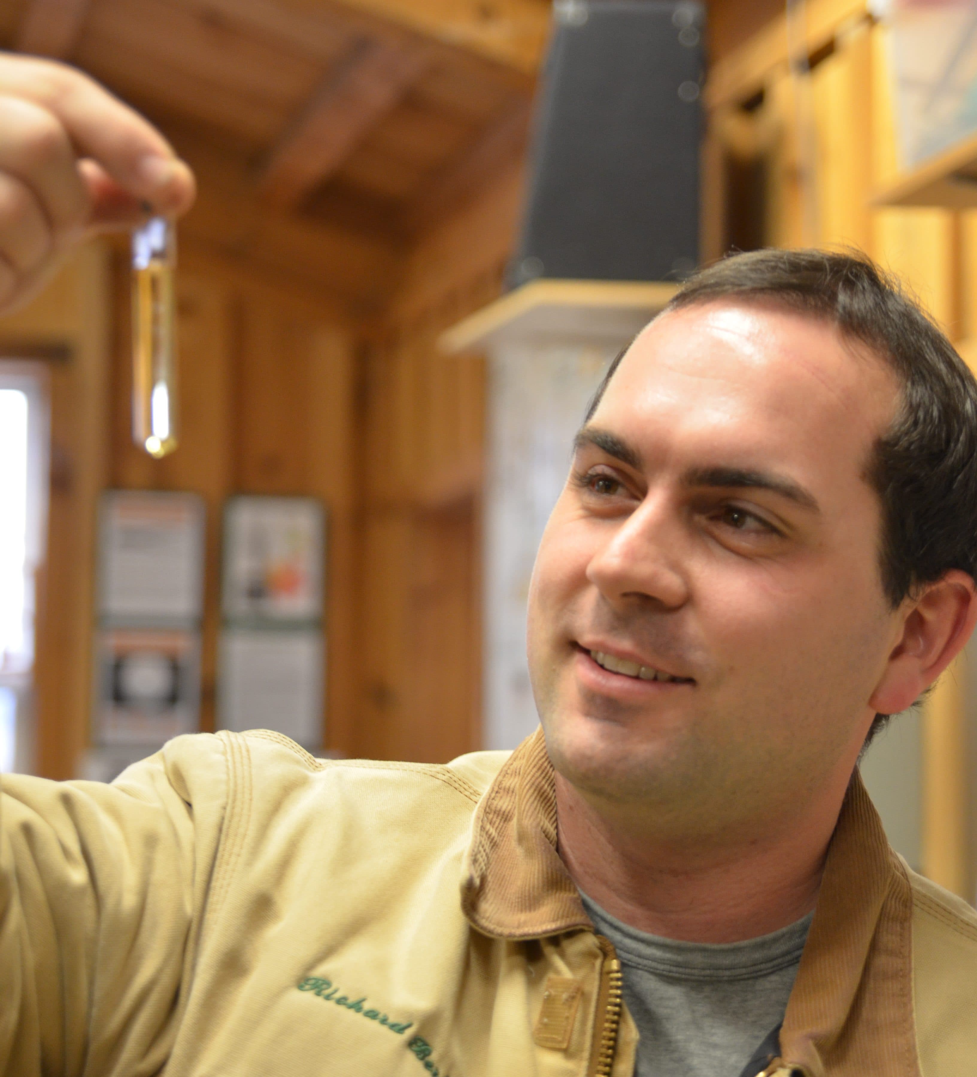Richard Bering, in a jacket embroidered with his name, smiles as he holds a small vial of maple syrup up to the light