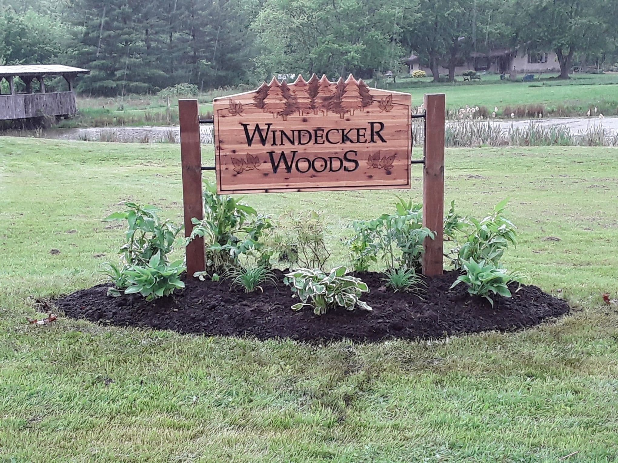 A carved cedar Windecker Woods sign planted with hostas, with the pond and covered bridge behind